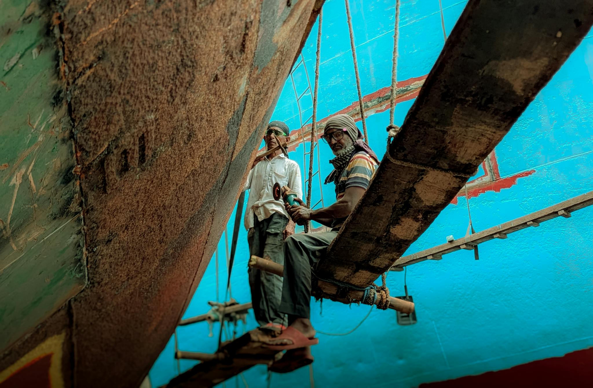 Two men work on painting a large ship in a shipyard, suspended on a plank.